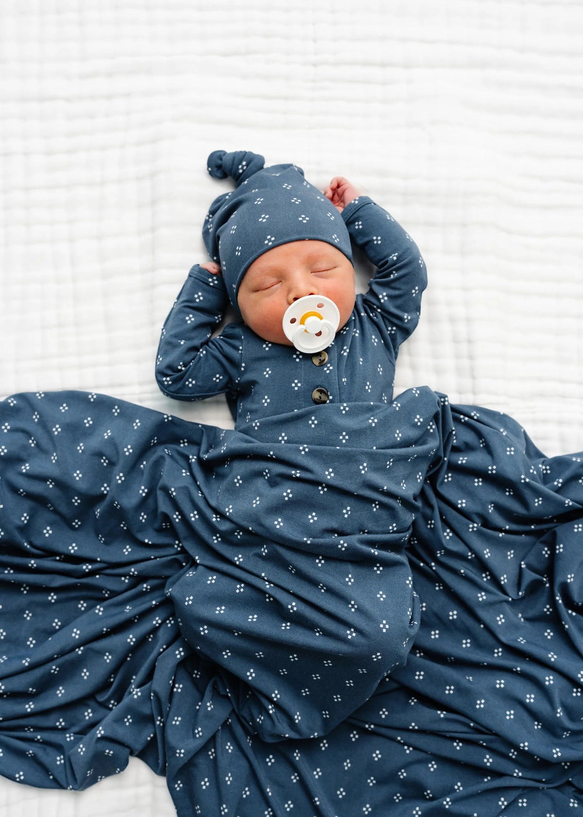 Newborn baby wrapped in a blue swaddle with a matching hat and pacifier, lying on a white textured surface.