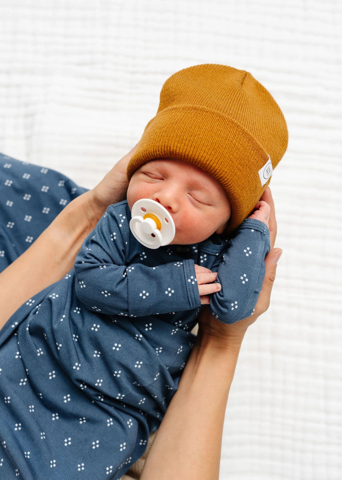 Newborn baby wearing a mustard hat and blue outfit with a white background