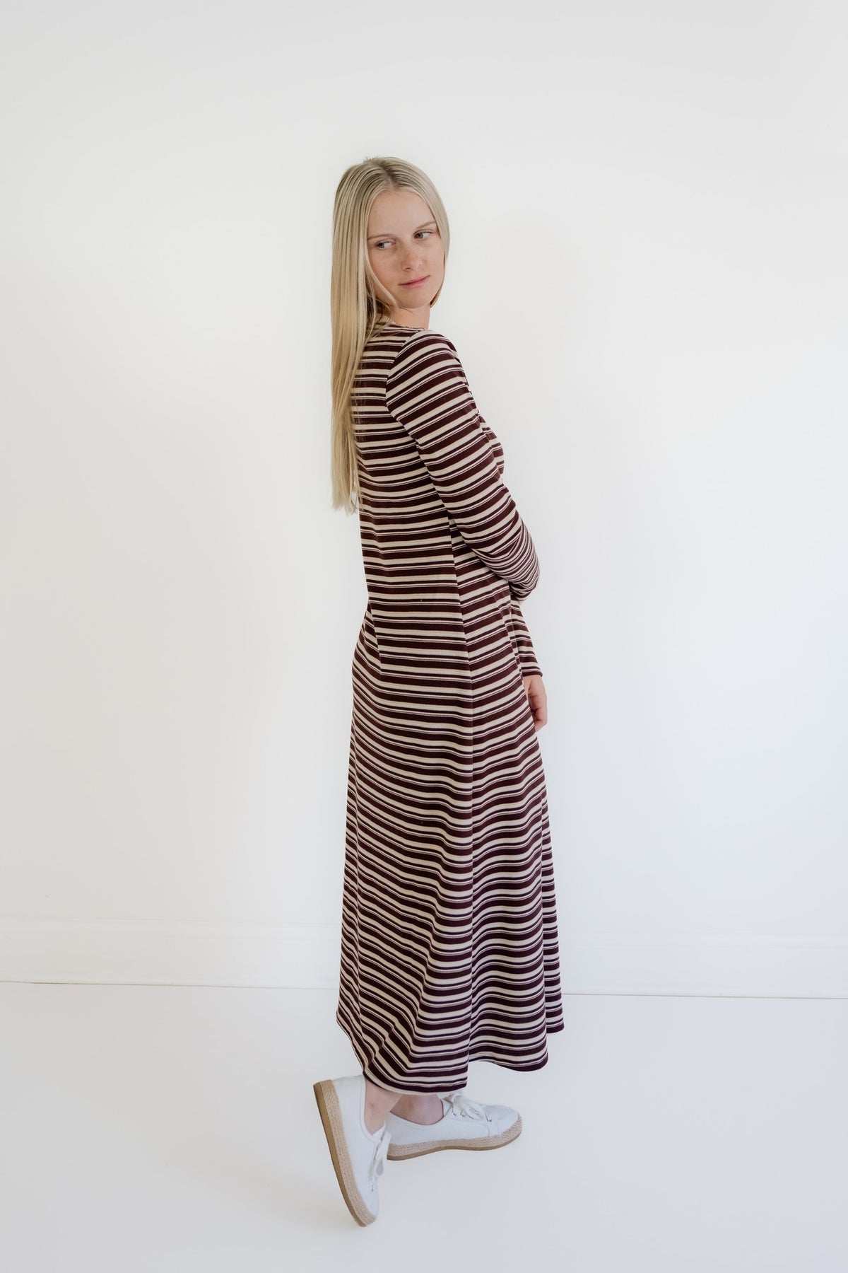 A woman with long blonde hair stands in profile wearing a long-sleeved maxi dress with horizontal brown, beige, and white stripes, paired with white slip-on shoes, against a plain white studio background.