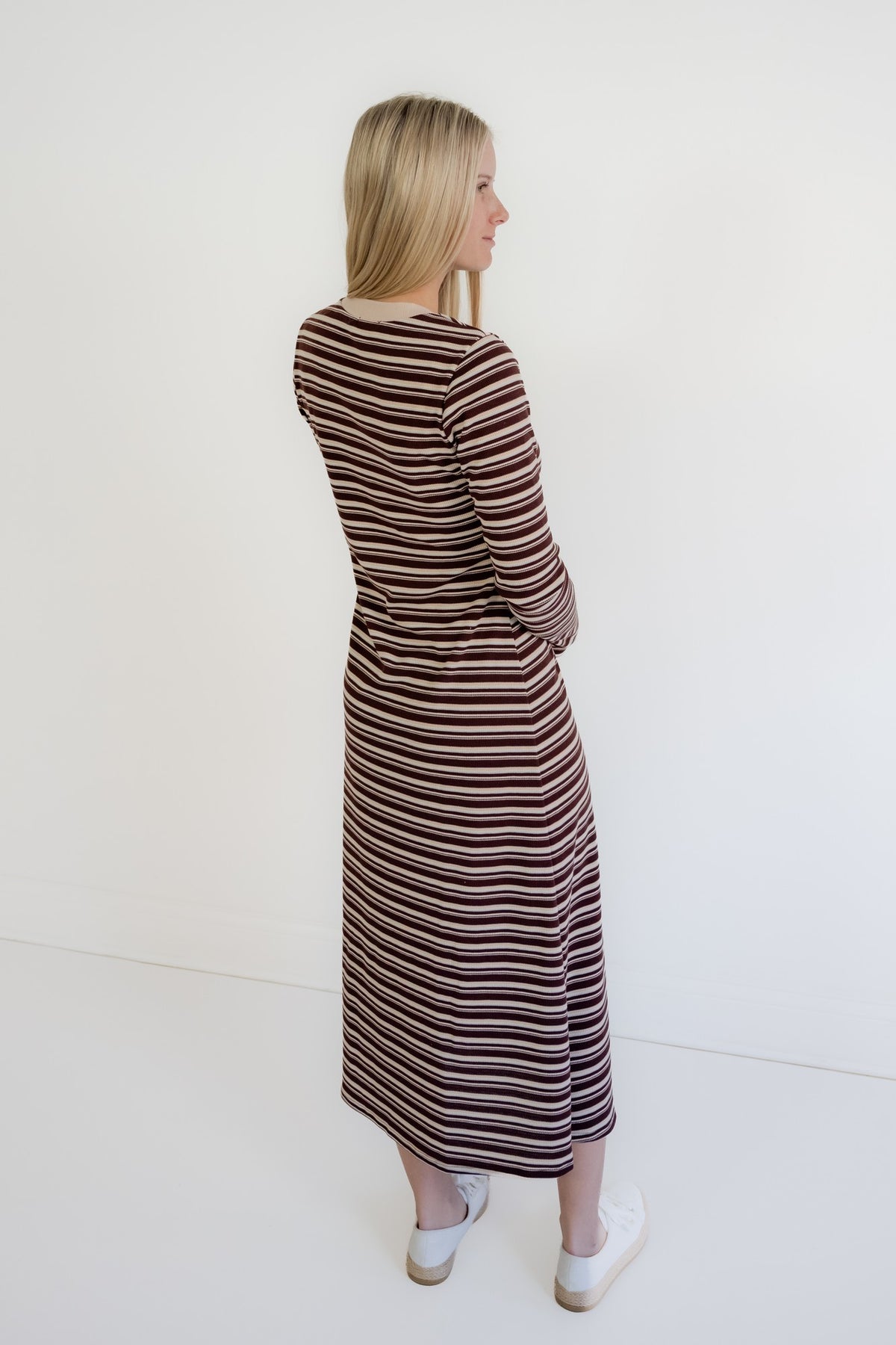 A woman with blonde hair is shown from the back wearing a long-sleeved midi dress with thin horizontal stripes in brown, beige, and white. She is standing against a plain white wall.