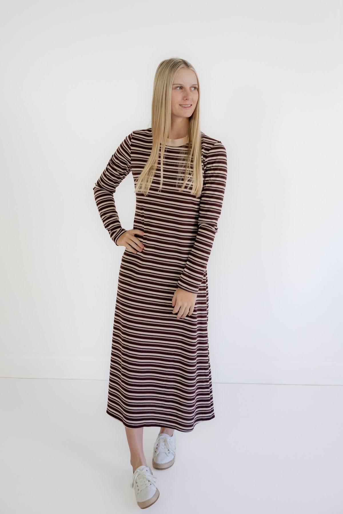 A woman modeling a long-sleeve midi dress with horizontal brown and beige stripes, paired with white sneakers, against a plain white studio background.