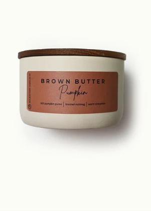 A candle in a white ceramic jar with a brown wooden lid and a terracotta-colored label that reads 'BROWN BUTTER Pumpkin'.