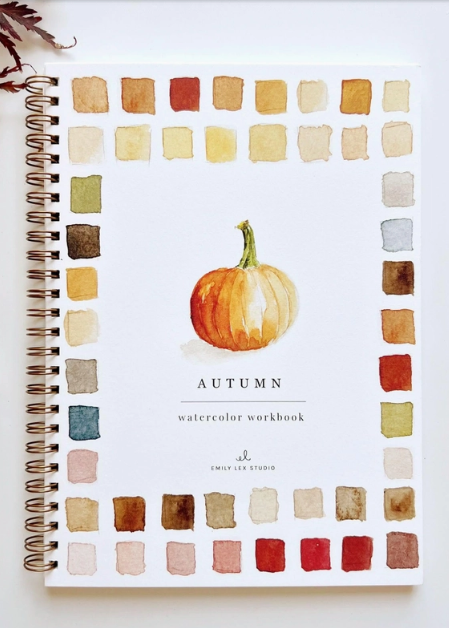 A spiral-bound watercolor workbook titled 'AUTUMN' with a watercolor pumpkin illustration, surrounded by a border of watercolor color swatches. The workbook is photographed on a white surface with a paintbrush, paint tube, and dried leaves as styled props.