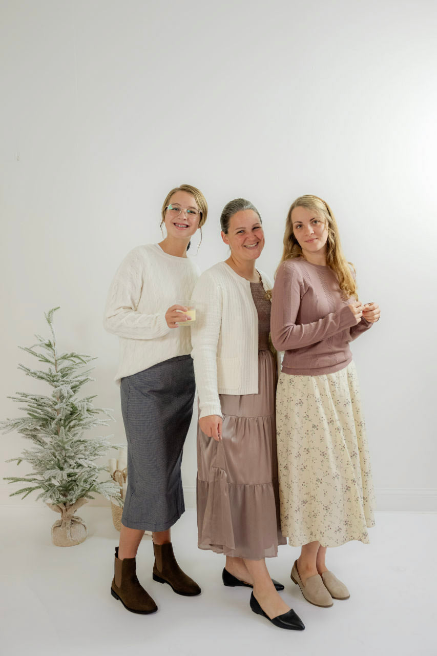 Three women in modest skirts and sweaters posing indoors by a small frosted tree, Salt and Honey boutique style