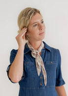 A woman modeling a blue denim midi dress with short sleeves, a button-up front, and a matching neck scarf, standing against a plain white studio background.