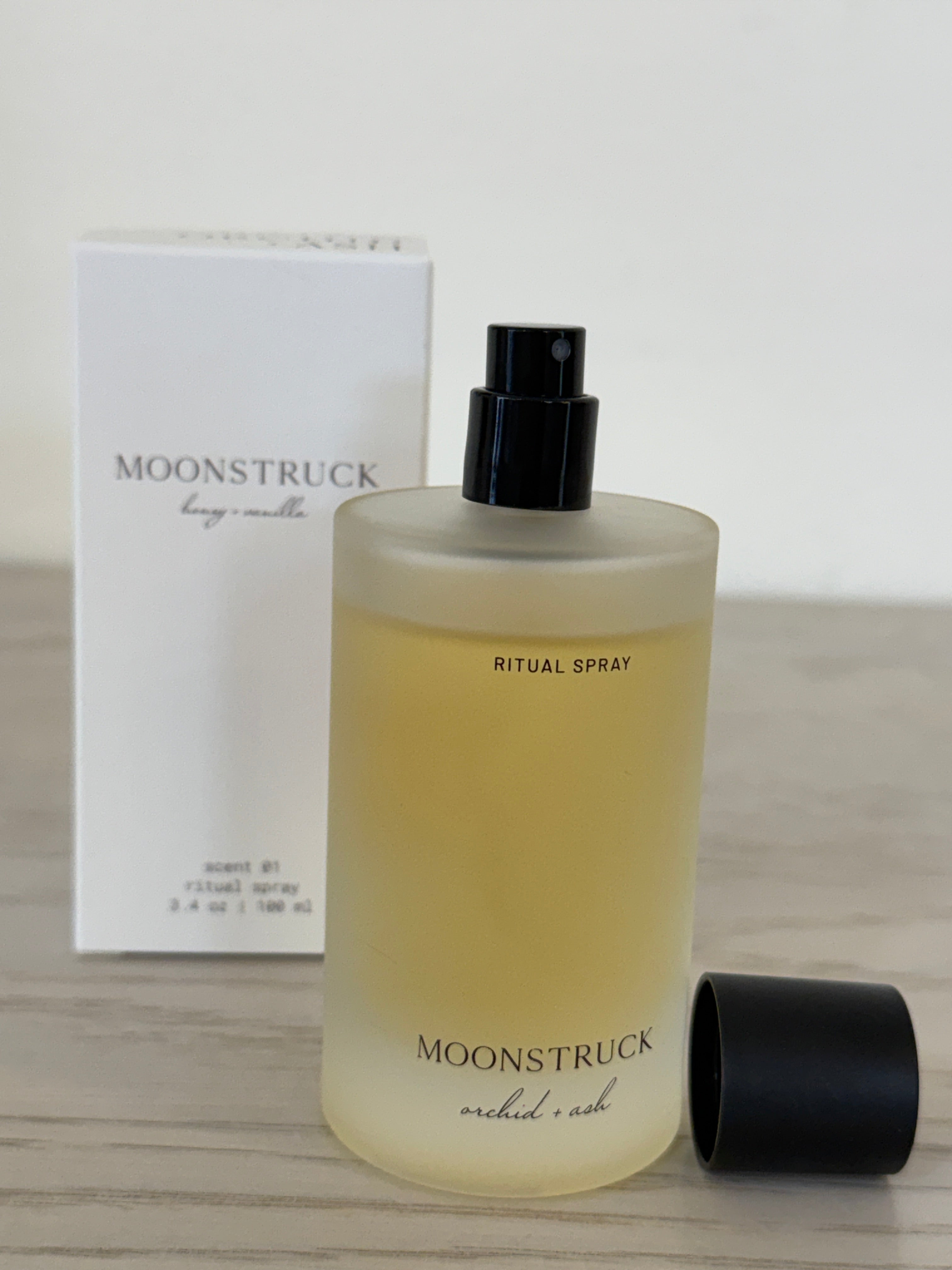 A bottle of Moonstruck Ritual Spray with a frosted glass body showing pale yellow liquid, a black spray top, and a black cap placed beside it, with its white product box behind on a light wood surface.