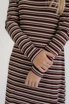 A close-up of a person wearing a long-sleeve knit dress with horizontal dark brown and beige stripes, with their hands folded over in front of the chest.