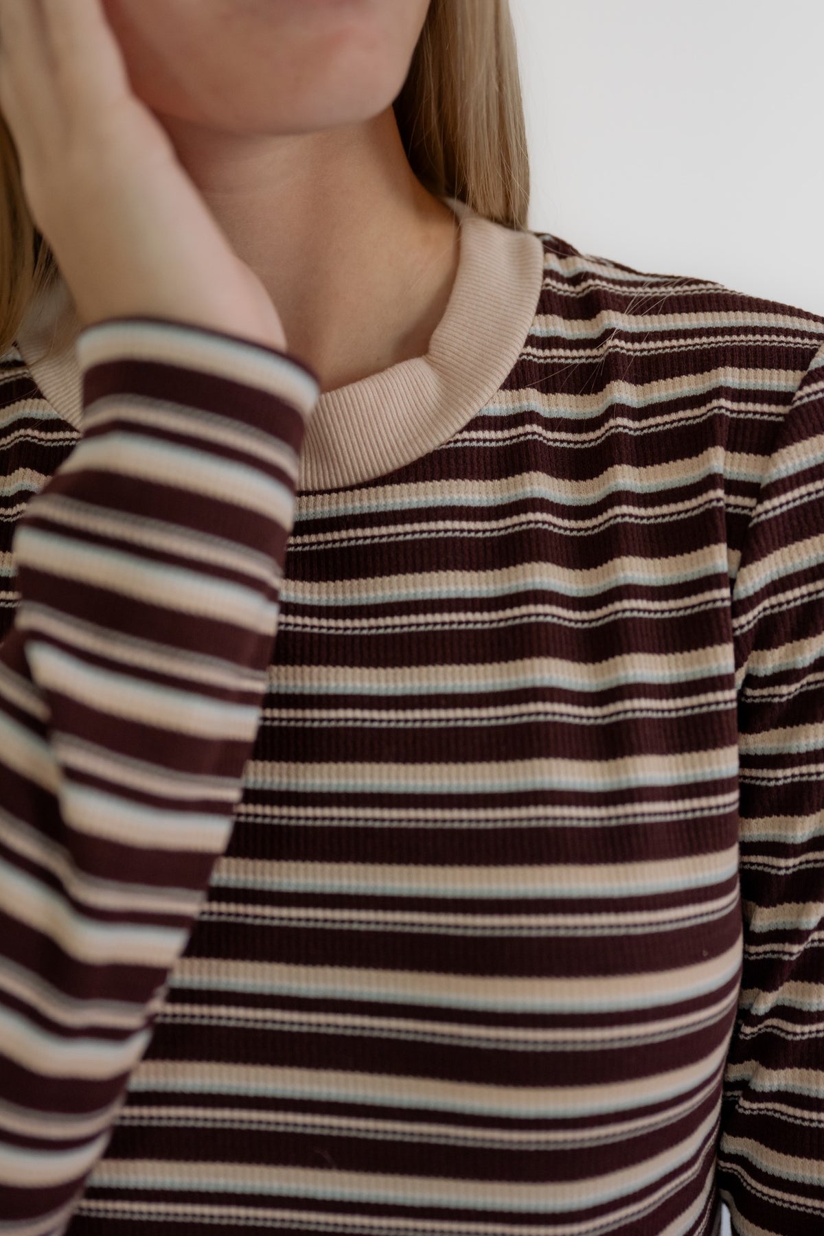 Close-up of a person wearing a ribbed long-sleeve top with horizontal stripes in dark brown, beige, and white, featuring a solid beige crew neck.