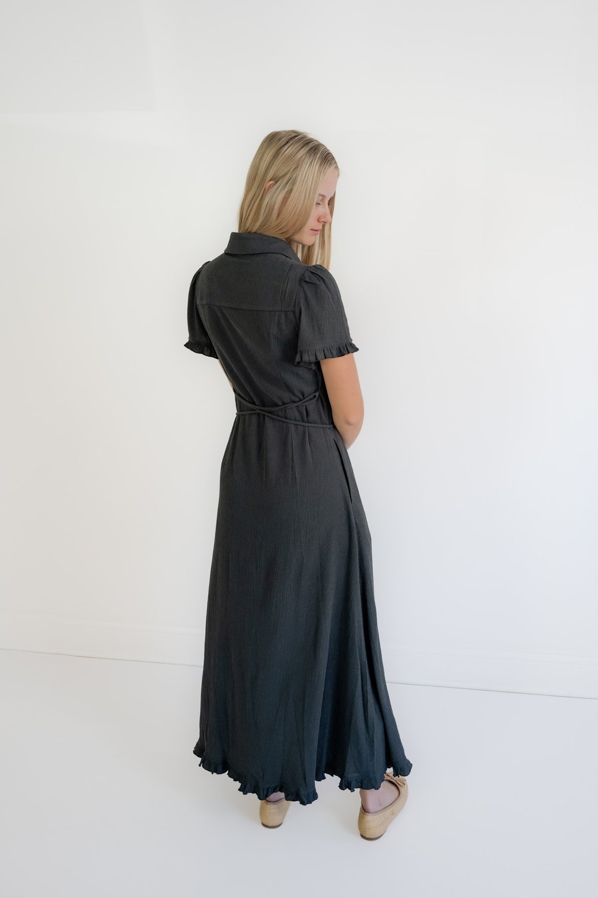 A woman with blonde hair is shown from behind wearing a dark charcoal gray short-sleeve collared dress with a tie waist, standing against a plain white studio background.