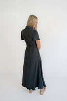 A woman with blonde hair is shown from behind wearing a dark charcoal gray short-sleeve collared dress with a tie waist, standing against a plain white studio background.