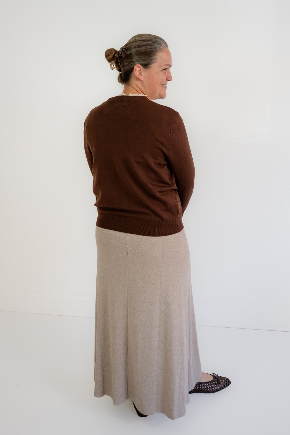 A woman shown from the back wearing a dark brown long-sleeve knit top and a light beige maxi skirt, standing against a plain white studio background.