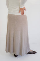 A woman wearing a long, flowing A-line maxi skirt in a light taupe-beige color, paired with a white long-sleeve top and black woven flat shoes, shown from the waist down against a white studio background.