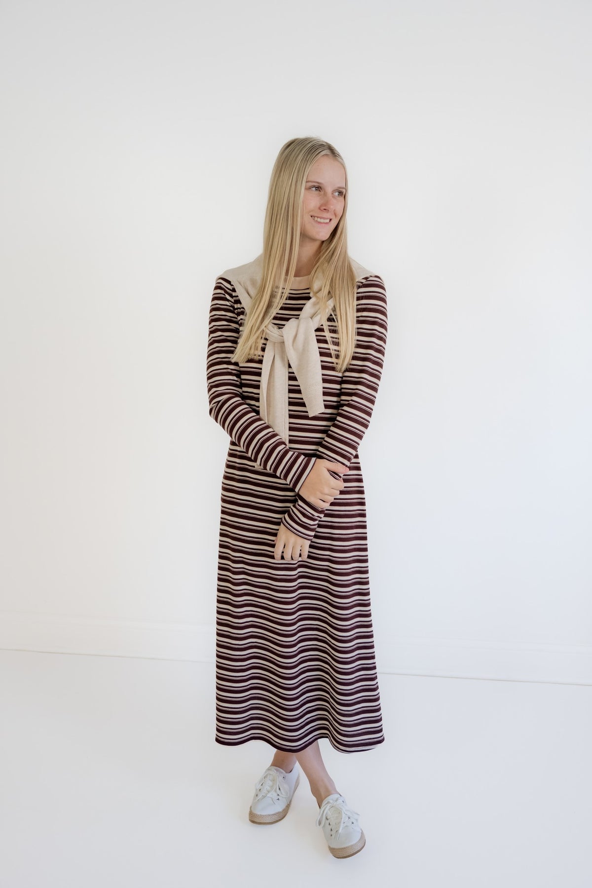 A woman modeling a long-sleeve knit midi dress with horizontal stripes in brown, burgundy, and cream, featuring a cream bow at the neckline. She is wearing white sneakers and standing against a plain white studio background.