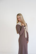 A blonde woman wearing a long-sleeve, knee-length striped dress in brown and beige, styled with a cream knit scarf, standing against a plain white wall.