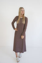 A woman modeling a long-sleeve midi dress with horizontal brown and beige stripes, paired with white sneakers, against a plain white studio background.