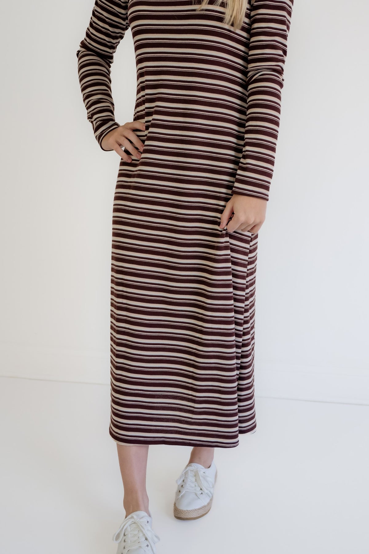 A woman wearing a long-sleeve midi dress with horizontal stripes in dark brown, burgundy, and beige, paired with white sneakers, against a plain white background.