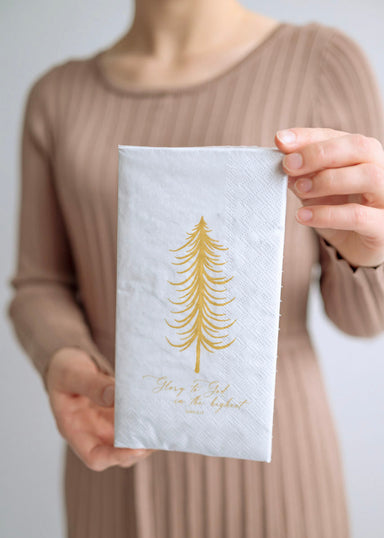 A person holding a white paper napkin with a gold tree print and the text 'Glory to God' on it.