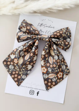 A fabric hair bow with a brown, beige, and white floral and leaf pattern, displayed on a white branded card.