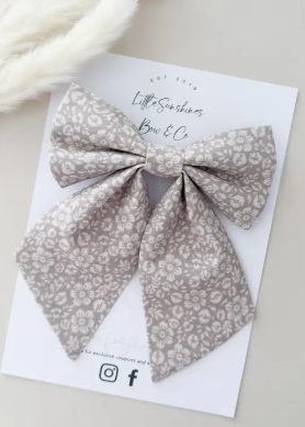 A large fabric hair bow with a beige and white floral pattern, displayed on a white branded card that reads 'Little Sunshines Bow & Co'.