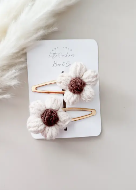 Two gold-tone hair clips decorated with white crocheted daisy flowers and dark brown centers, displayed on a white backing card with a soft beige background and dried white grass.
