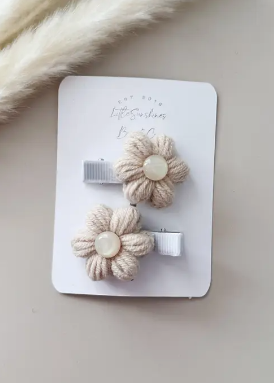 Two small flower-shaped hair clips in a cream textured fabric with pearl-like centers, attached to a white display card. The card is photographed on a light beige surface with a soft white faux-fur prop in the top left corner.