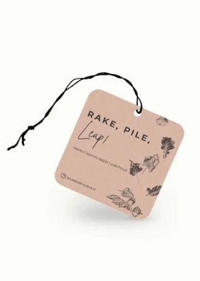 A square kraft paper tag with rounded corners, featuring the text 'RAKE, PILE, LEAP!' and botanical illustrations, suspended by a black string on a clean white background.