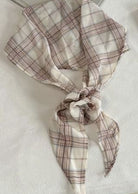 A lightweight square scarf with a beige and brown plaid pattern, tied in a knot and draped on a white fabric surface.