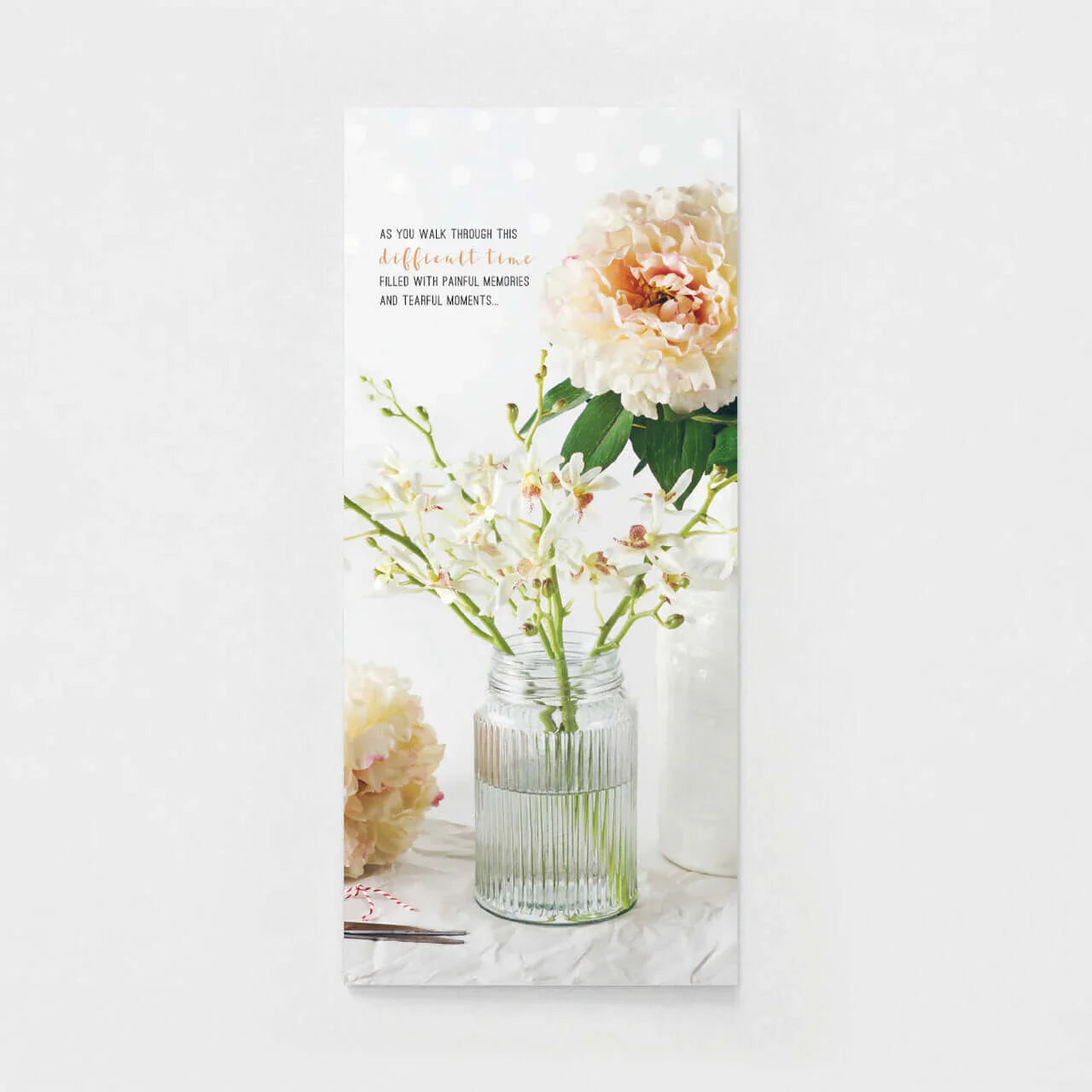 Sympathy card with soft pink peonies and white flowers in a glass vase on a white table.